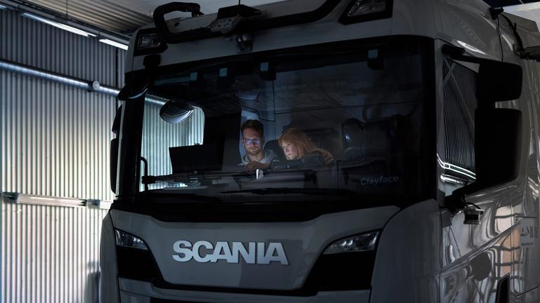 Two people sitting in the cab of a white Scania truck viewed through the windshield in an industrial warehouse setting.