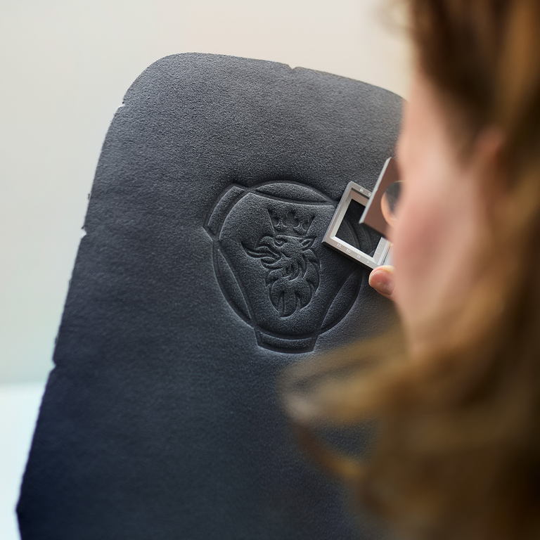 Person inspecting a black material panel embossed with the Scania griffin logo, using a small measuring tool for precision.