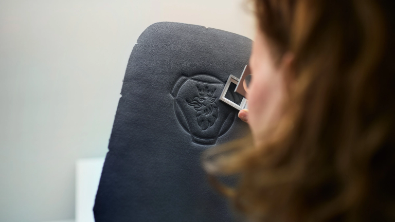 Person inspecting a black material panel embossed with the Scania griffin logo, using a small measuring tool for precision.