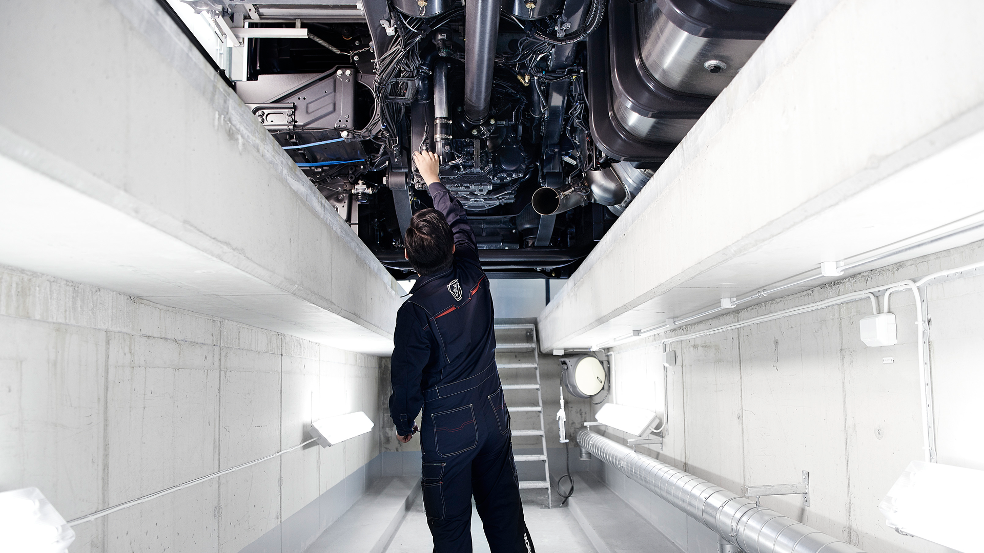 Mechanic working underneath a Scania truck in a brightly lit service bay.
