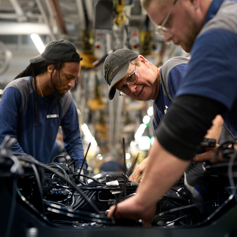 Three Scania workers assembling machinery and wiring on the factory floor.
