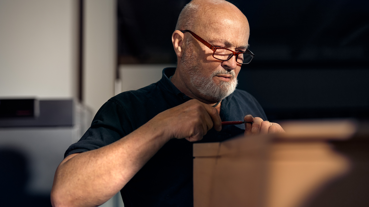 Man with glasses and a white beard concentrating while working with his hands on a wooden object, holding a red-handled tool.