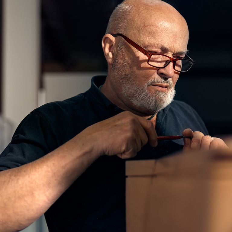 Man with glasses and a white beard concentrating while working with his hands on a wooden object, holding a red-handled tool.