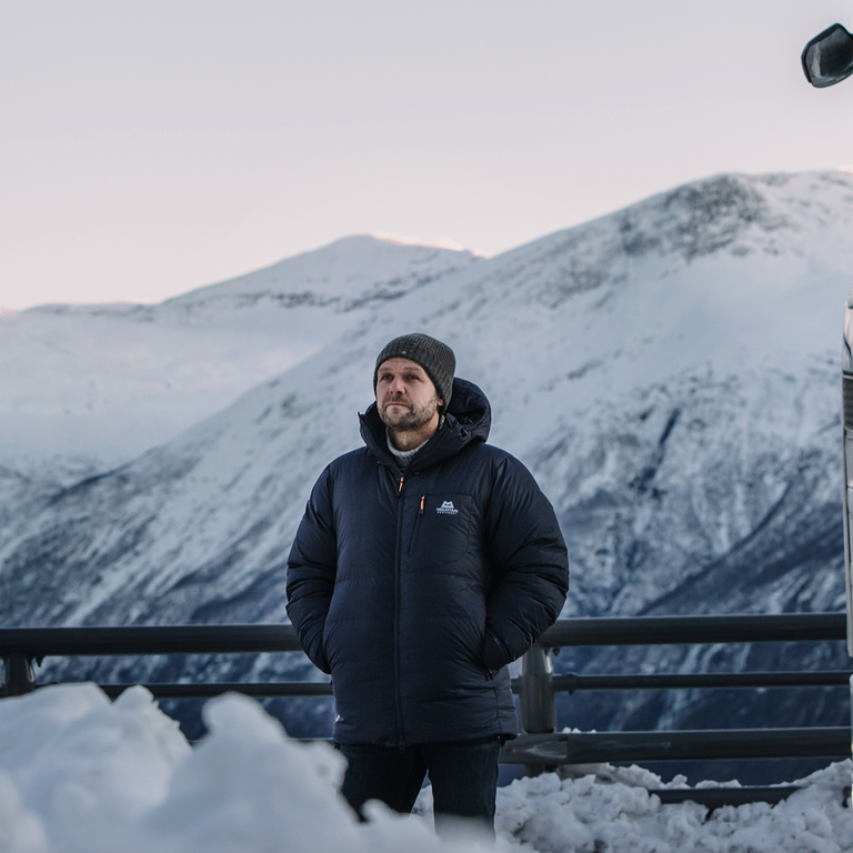 Man in a winter jacket and beanie standing beside a white Scania truck, set against a snowy mountain landscape in cold weather conditions.