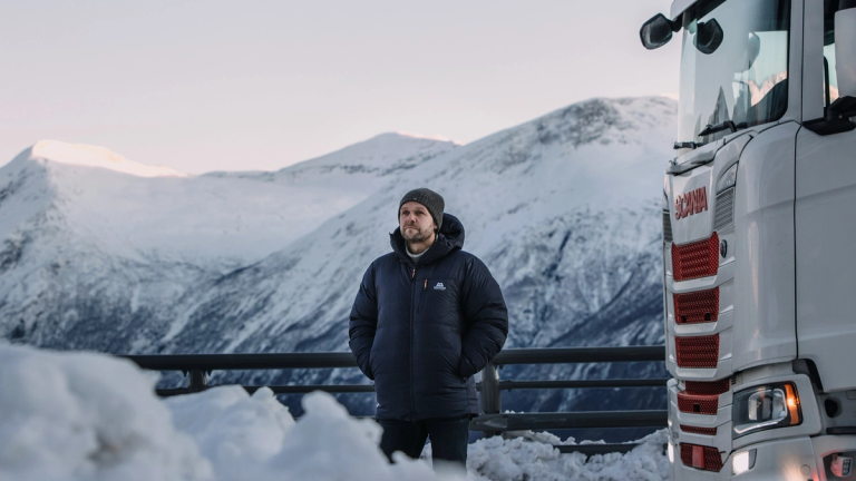 Man in a winter jacket and beanie standing beside a white Scania truck, set against a snowy mountain landscape in cold weather conditions.