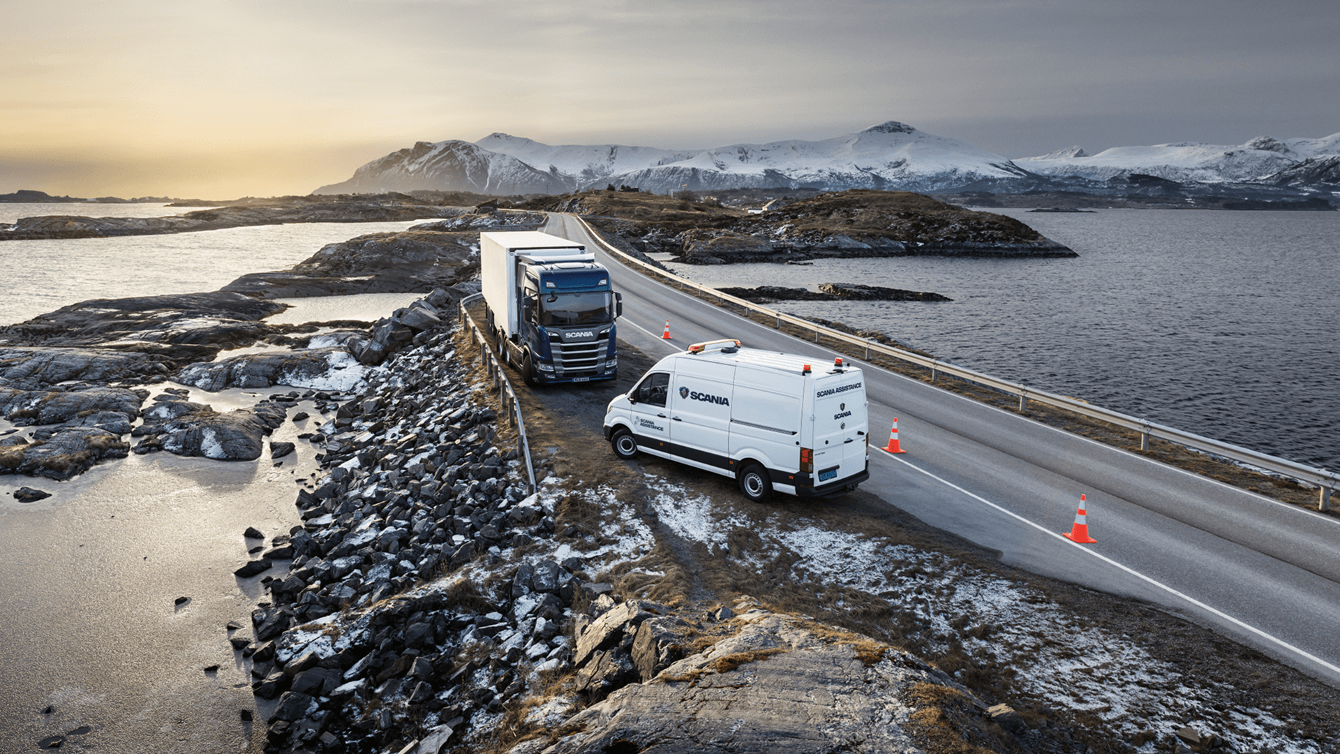 Scania service van assisting a broken-down truck on a winding coastal road, with a backdrop of snow-covered mountains at sunrise.