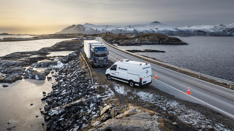 Scania service van assisting a broken-down truck on a winding coastal road, with a backdrop of snow-covered mountains at sunrise.
