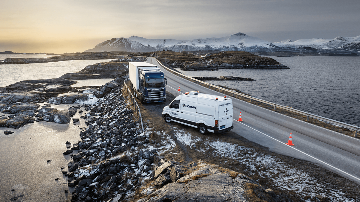 Scania service van assisting a broken-down truck on a winding coastal road, with a backdrop of snow-covered mountains at sunrise.