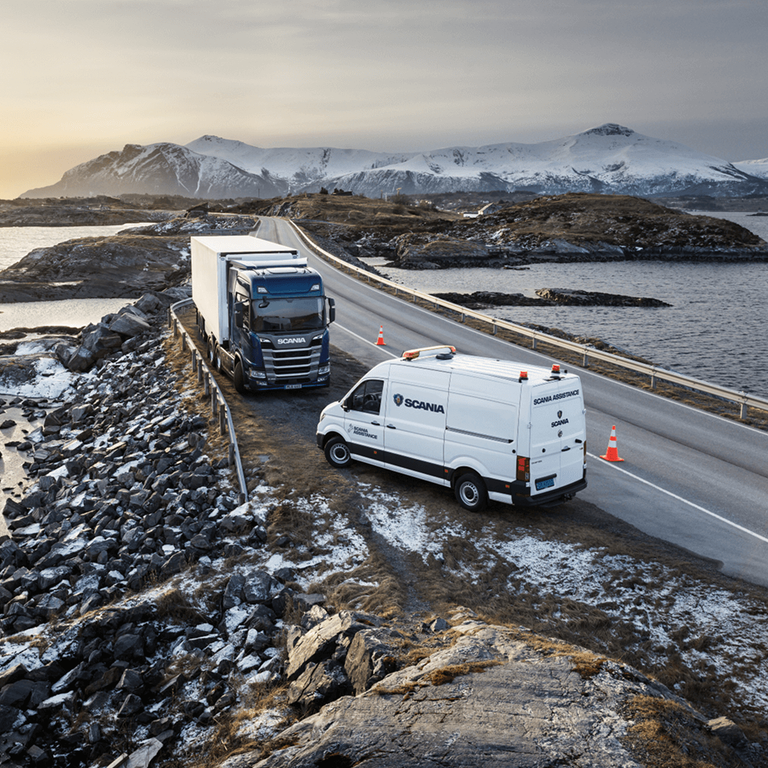 Scania service van assisting a broken-down truck on a winding coastal road, with a backdrop of snow-covered mountains at sunrise.