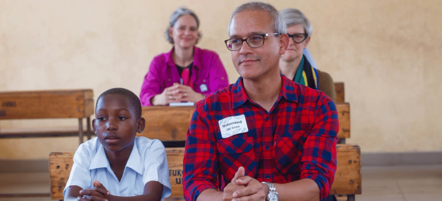 A man wearing glasses and a red plaid shirt sits at a wooden desk next to a young boy in a school uniform. Behind them, a few other adults sit attentively. The setting appears to be a classroom with wooden benches.