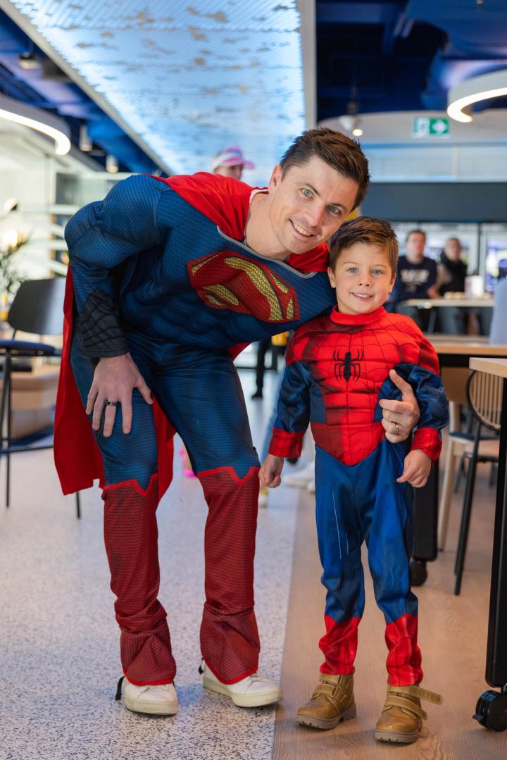 An adult and a child dressed in superhero costumes. The adult is wearing a Superman costume with a red cape, and the child is dressed as Spider-Man, standing next to the adult, smiling.