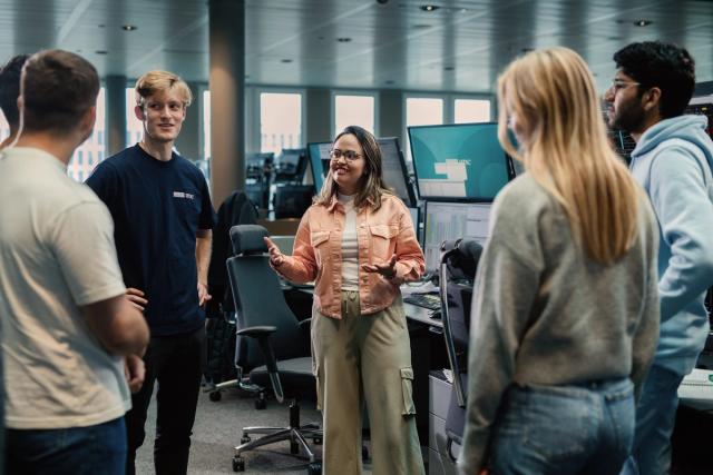 A group of young professionals standing in a circle in a modern office, engaging in a discussion. A woman in the center is speaking, gesturing with her hands, while the others listen attentively. Multiple computer monitors can be seen in the background.