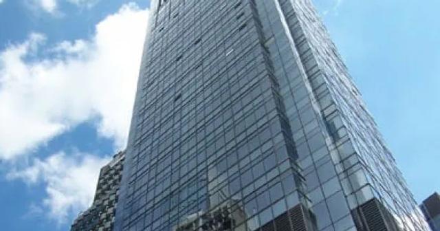 A tall glass skyscraper seen from a low-angle perspective against a bright blue sky with a few clouds. The building has reflective windows, giving it a sleek and modern appearance.