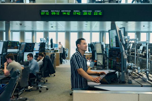 A modern office setting with multiple workers at desks, each using multiple computer monitors displaying financial data. A man stands in the foreground typing on a keyboard, with large screens in front of him. A digital world clock displaying various cities’ times hangs above the workspace.