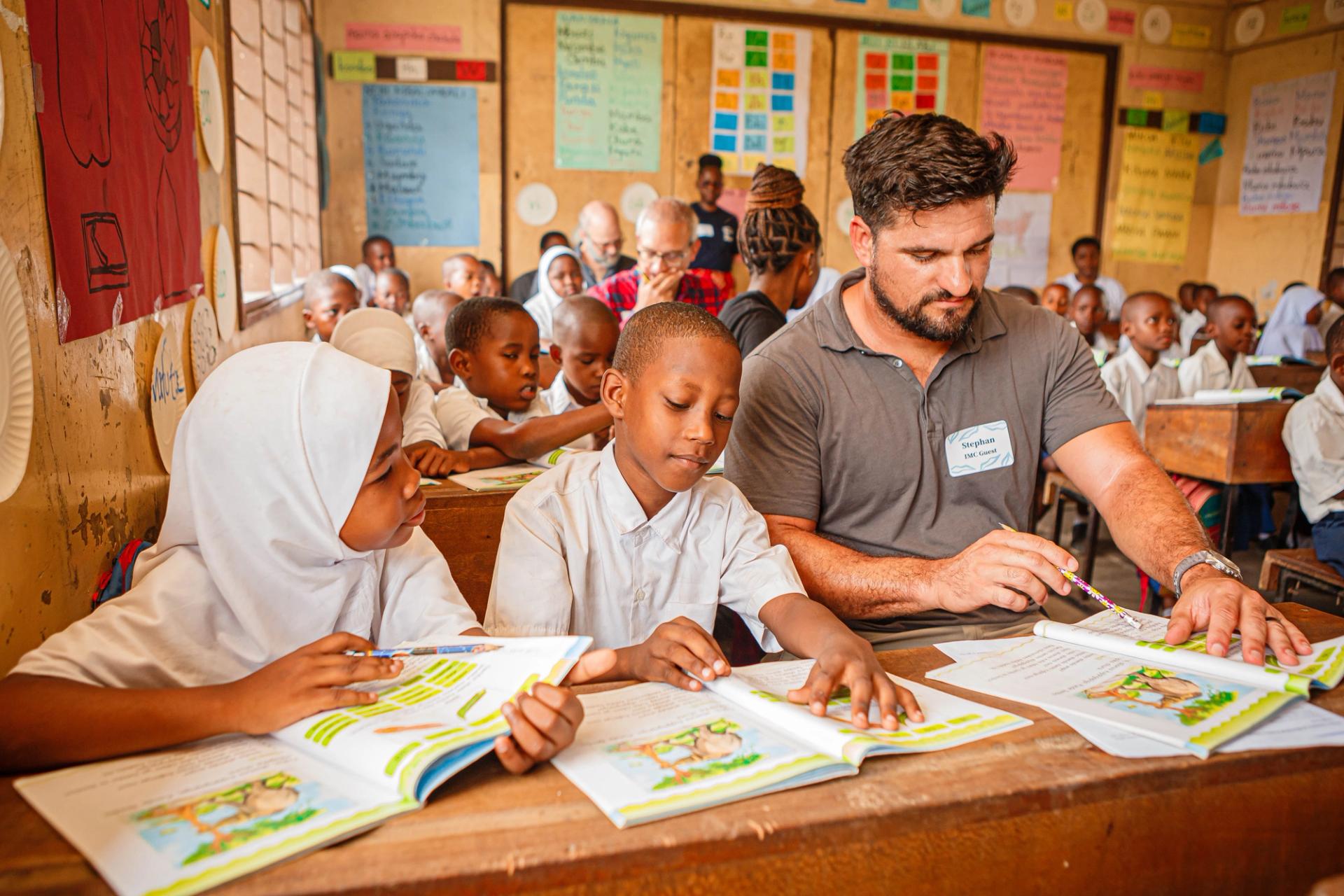 A classroom filled with young students in uniforms working on their lessons. A man seated at a desk with two students, helping them with their work. Colorful posters and educational materials adorn the walls of the classroom, and other children are focused on their books.