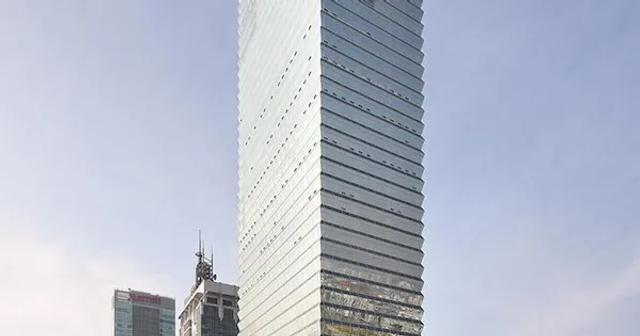 A tall, modern skyscraper with a glass facade, standing prominently against a clear sky. The building has a sleek, angular design, with nearby smaller buildings visible in the background.