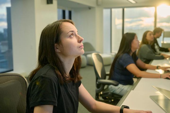 A young woman seated at a desk in an office, looking upward with a thoughtful expression. Behind her, other colleagues are working at their desks in a modern workspace with large windows letting in natural light from the setting sun.