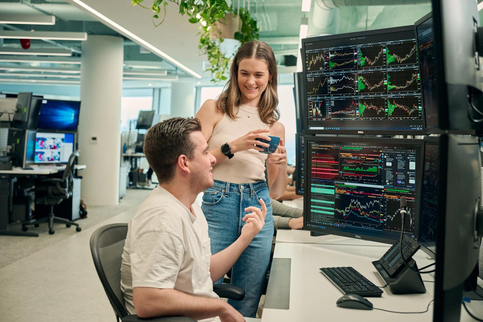 A man sitting at a desk with multiple computer monitors displaying financial charts and data, engaged in conversation with a woman standing next to him holding a coffee mug. Both are smiling, and the office environment is modern with plants hanging overhead.