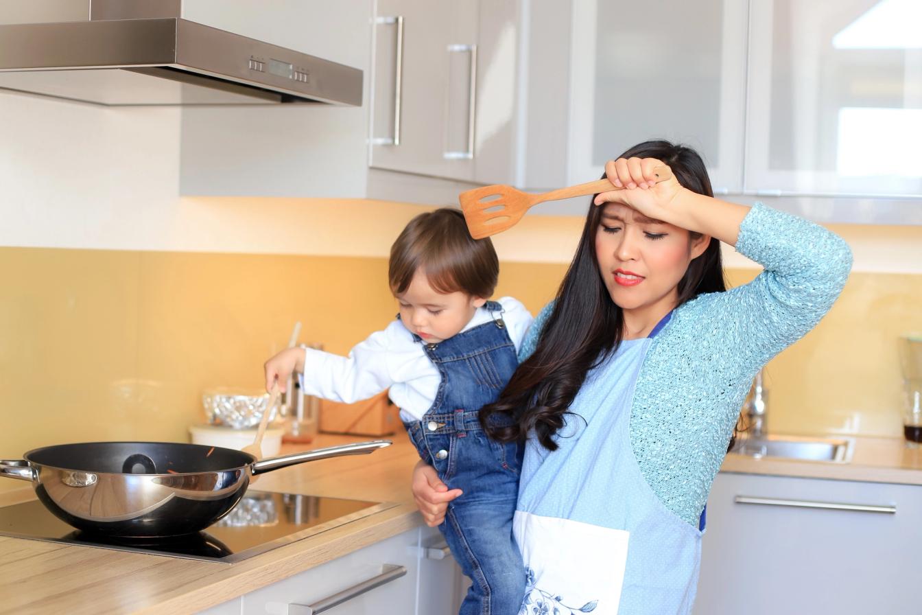 Ibu menggendong anak sambil memasak di dapur, menggambarkan kesibukan harian saat menyiapkan lauk MPASI