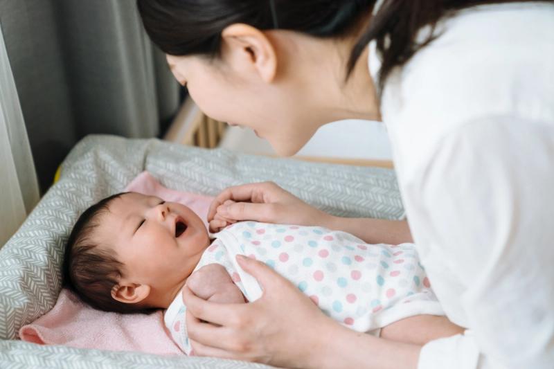 Ibu menenangkan bayi baru lahir yang menguap di meja ganti popok, menggambarkan perhatian orang tua dalam merawat kebutuhan bayi