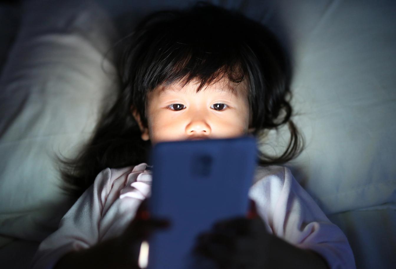 Anak perempuan berbaring di tempat tidur malam hari sambil menatap layar ponsel, menggambarkan rutinitas sebelum tidur anak