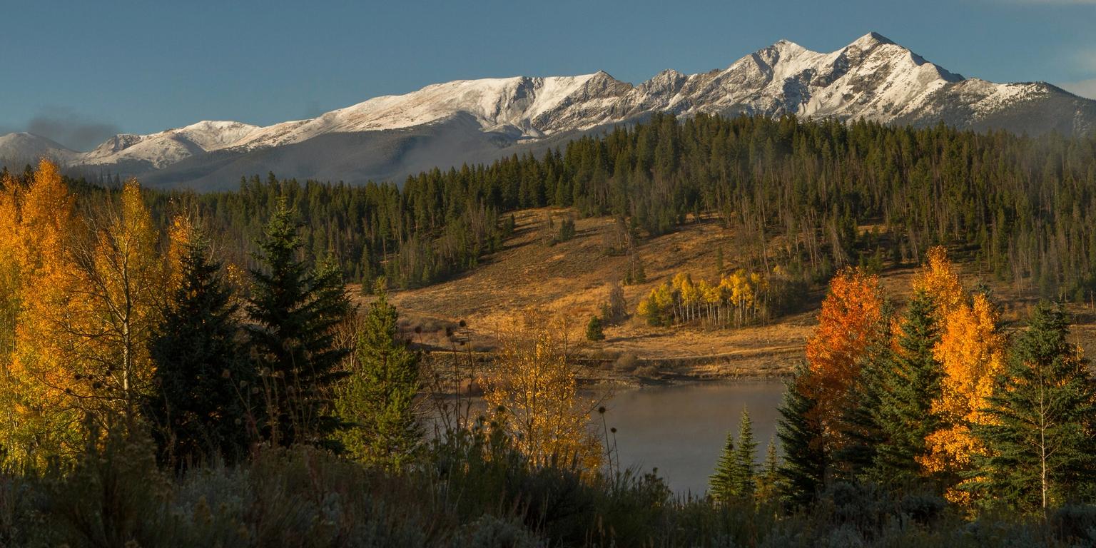 Snow-capped mountains overlook a lake surrounded by green hills and vibrant yellow and orange fall foliage.