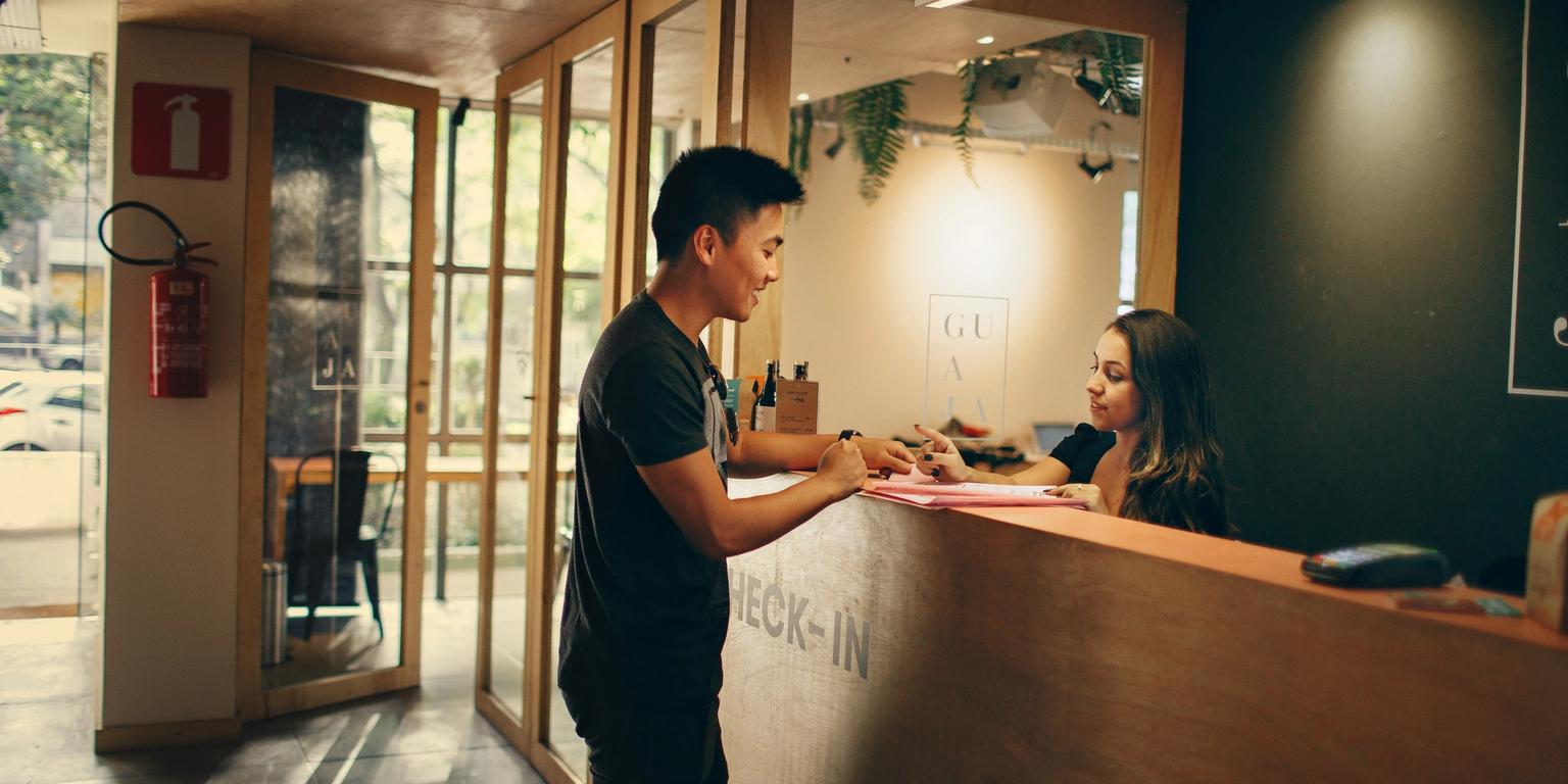A man stands at a check-in desk, interacting with a female receptionist.