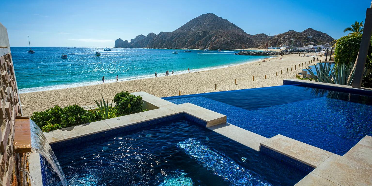 An infinity pool and hot tub overlook a vibrant beach with turquoise water, mountains, and boats.