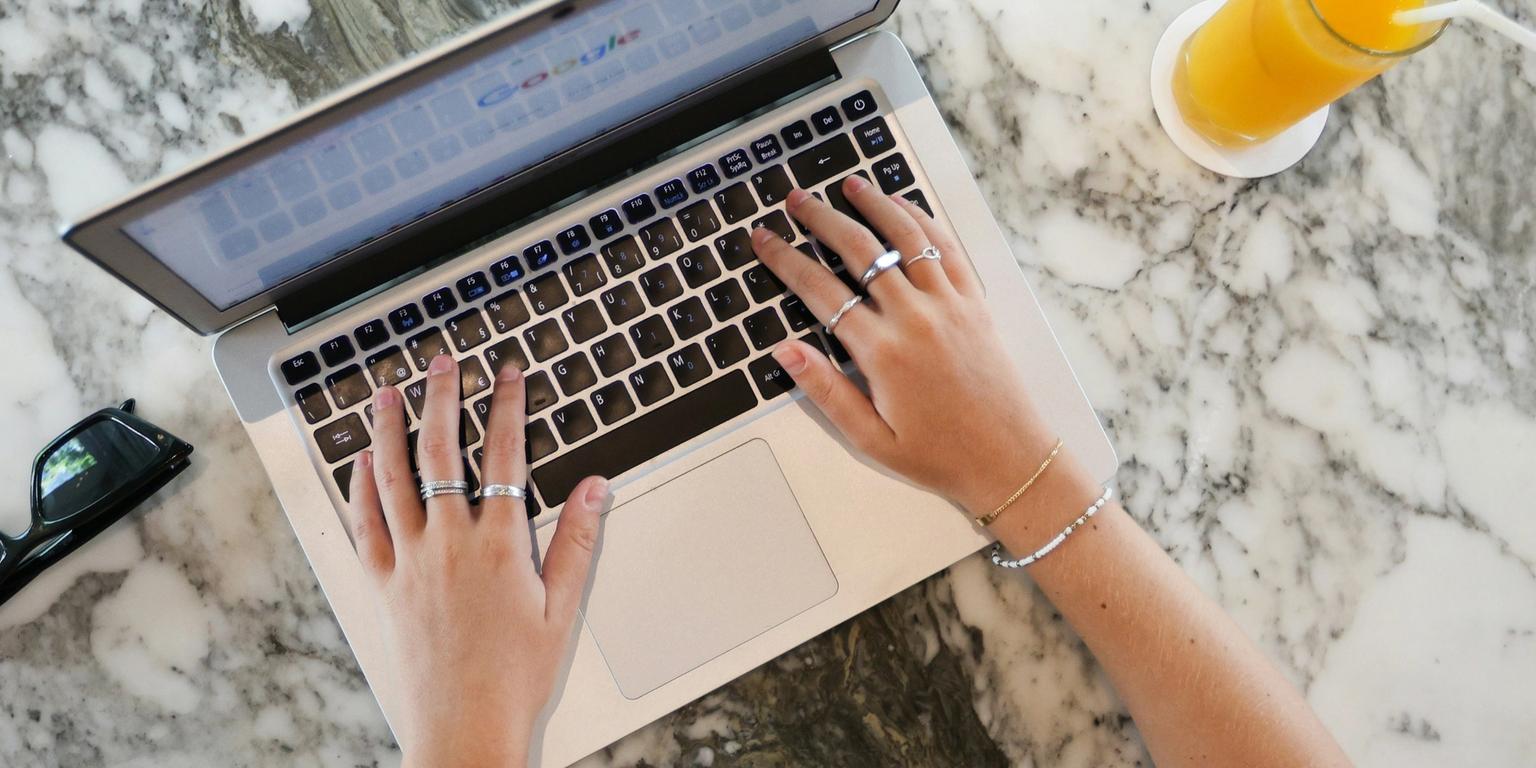 Overhead view of hands typing on a laptop, with orange juice and sunglasses on a marble table.