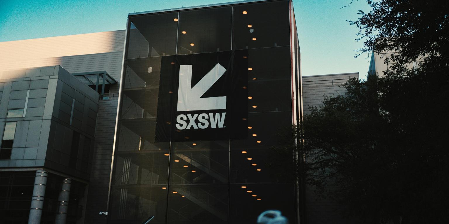 A modern glass building prominently displays a white SXSW logo and arrow.