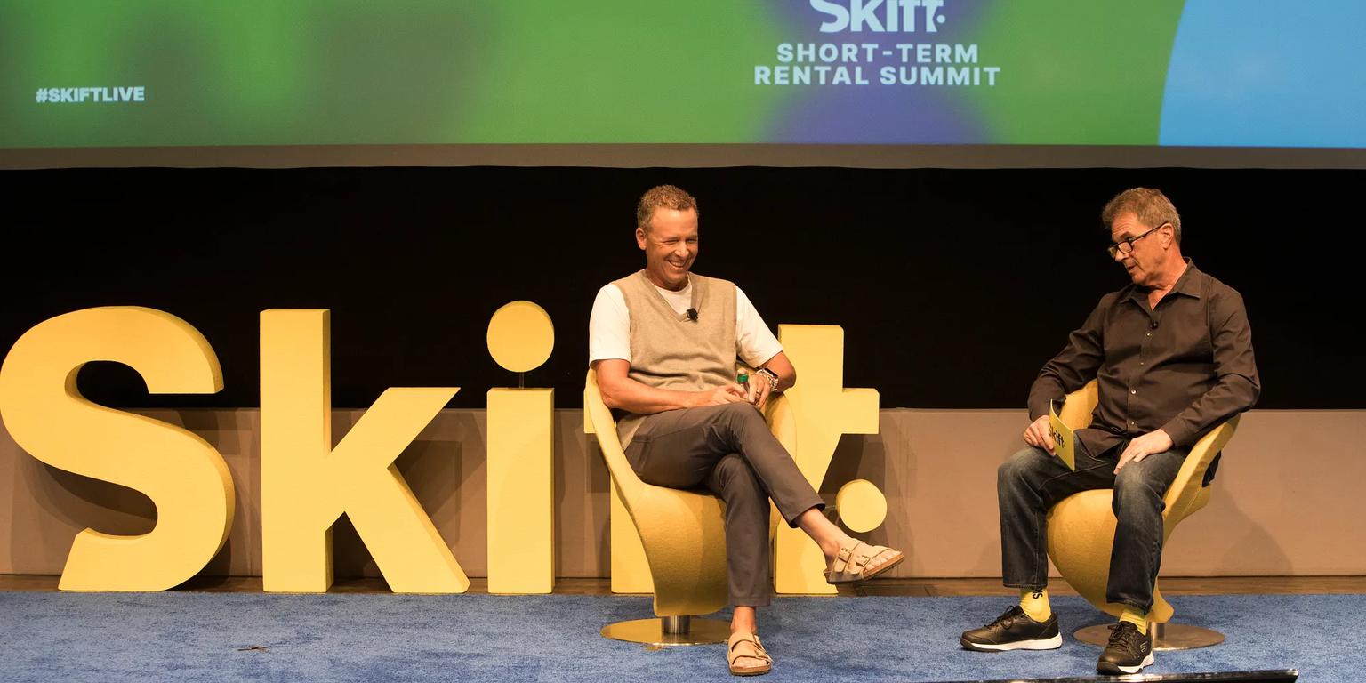 Two men converse on stage at the Skift Short-Term Rental Summit, flanked by large yellow "Skift" letters and event branding on a screen.