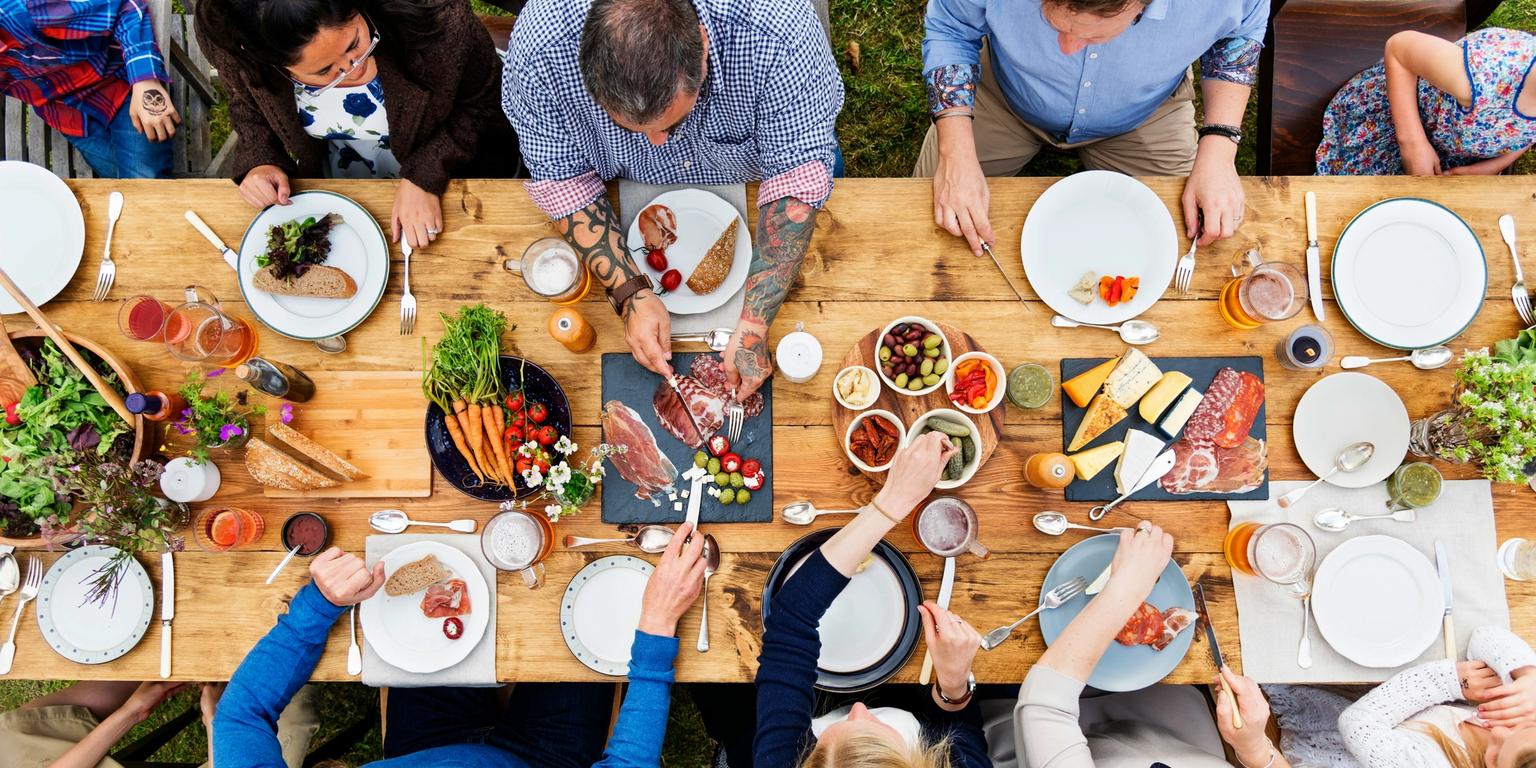 Overhead view of people sharing an outdoor meal at a long wooden table filled with food.