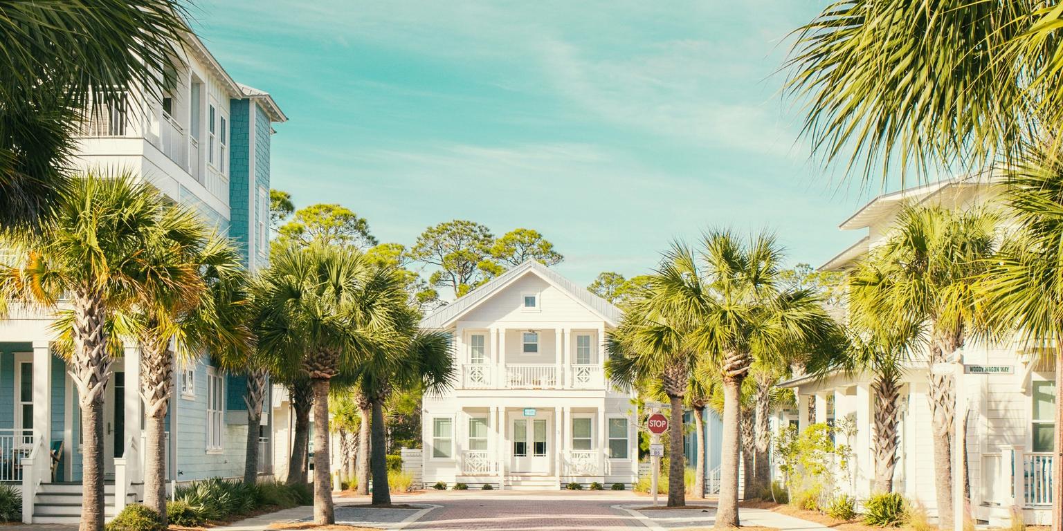 A brick-paved street lined with palm trees and coastal houses, leading to a white house at the end under a bright blue sky.