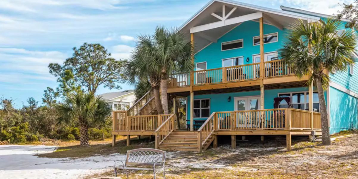A two-story turquoise beach house with wooden decks, palm trees, a fire pit, and a bench in a sandy yard.