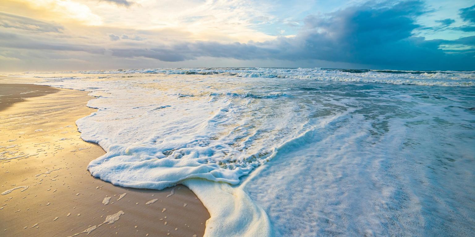 Ocean waves with white foam wash onto a sandy beach under a dramatic sky.