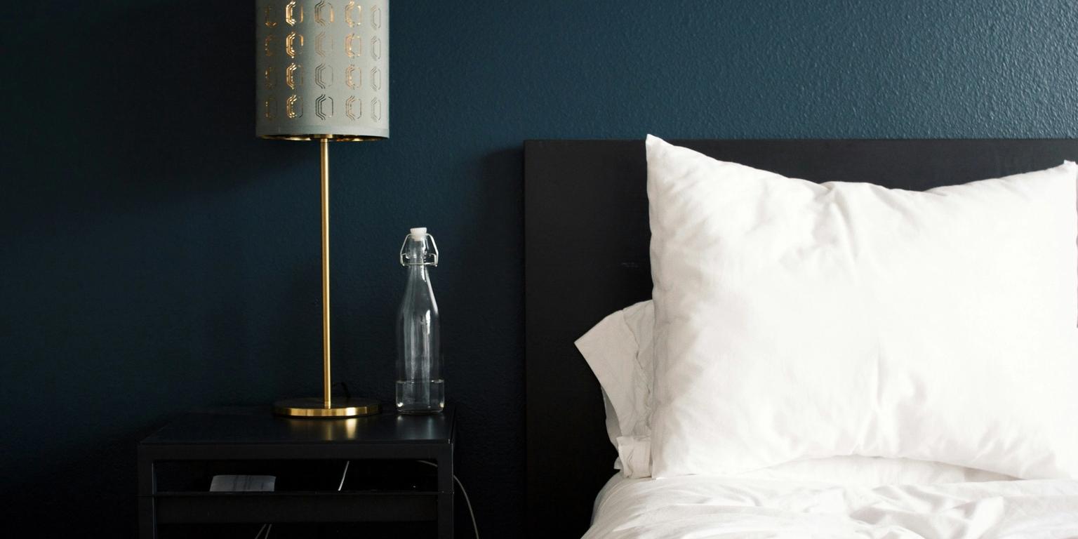 Brass lamp on a nightstand, white bed, and dark blue wall in a modern bedroom.