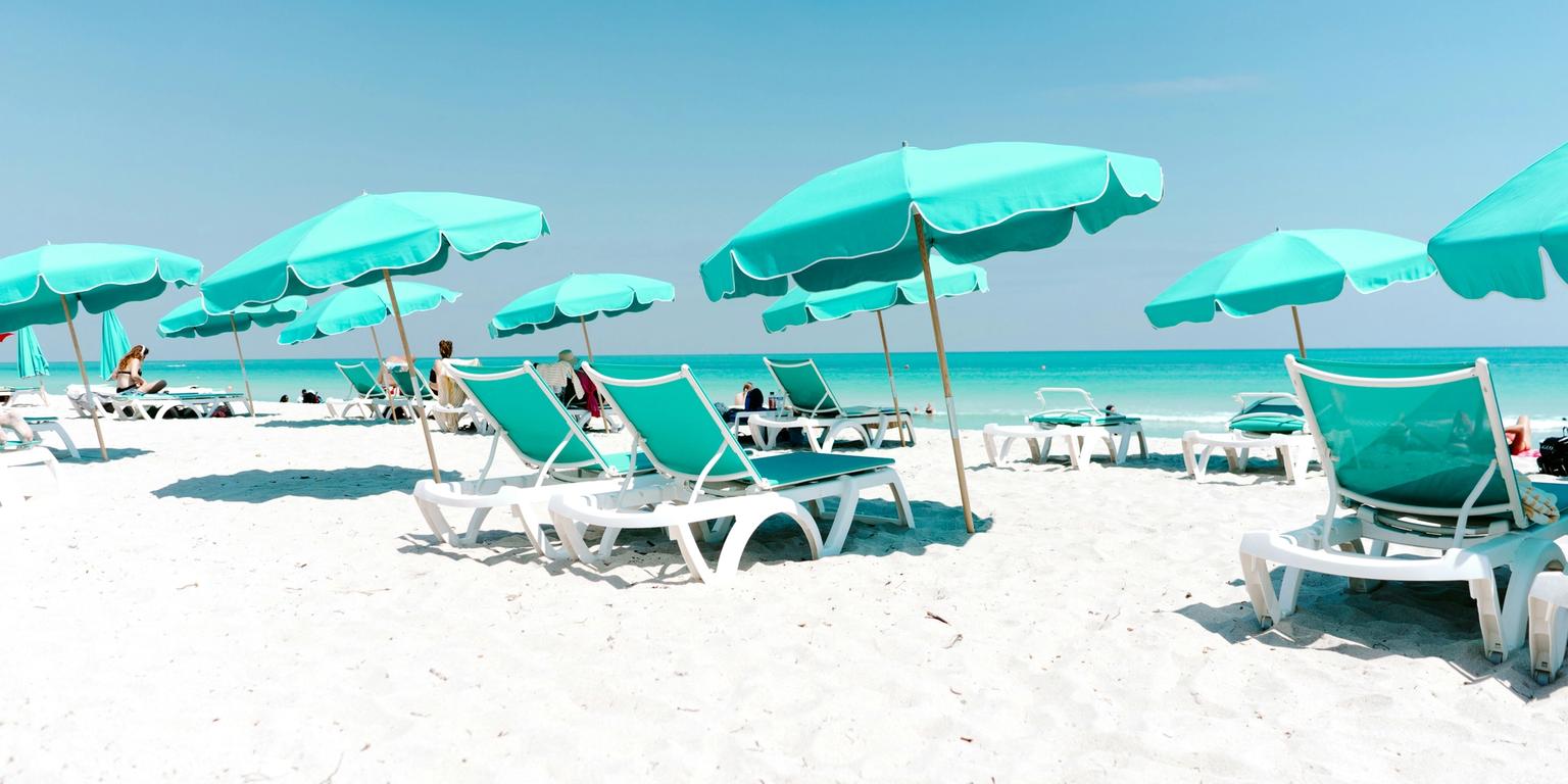 A white sand beach with rows of teal umbrellas and lounge chairs by the ocean.