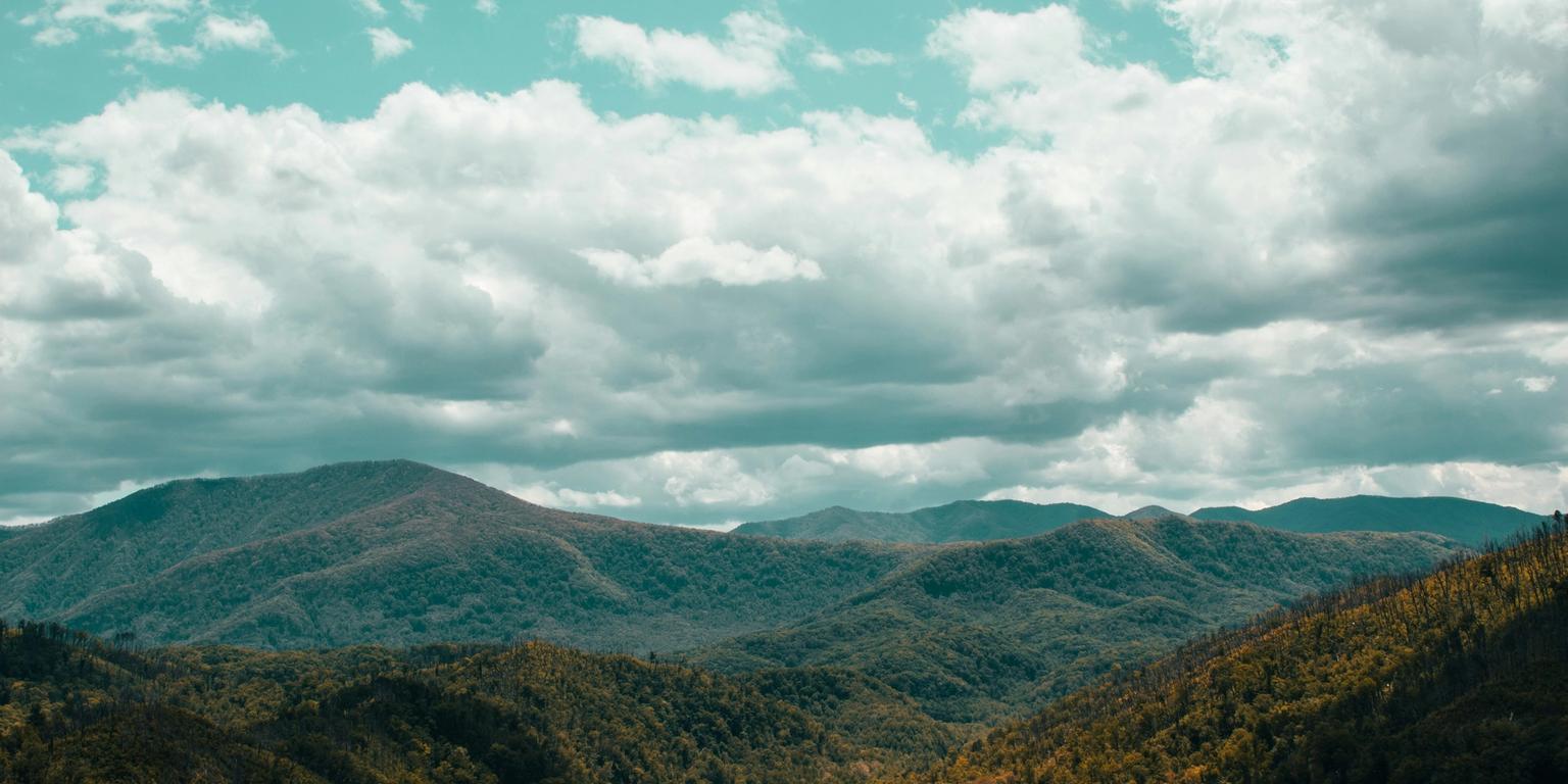 Forested mountains under a teal sky with fluffy white and grey clouds.