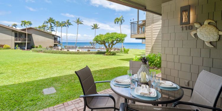Patio with a dining table set for four, overlooking a green lawn, palm trees, and the ocean.