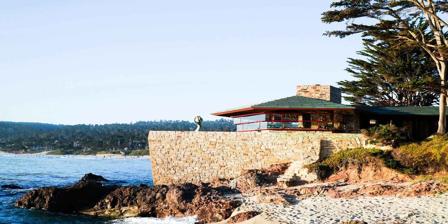 A stone house with a green roof stands on a rocky bluff overlooking a sandy beach and ocean.
