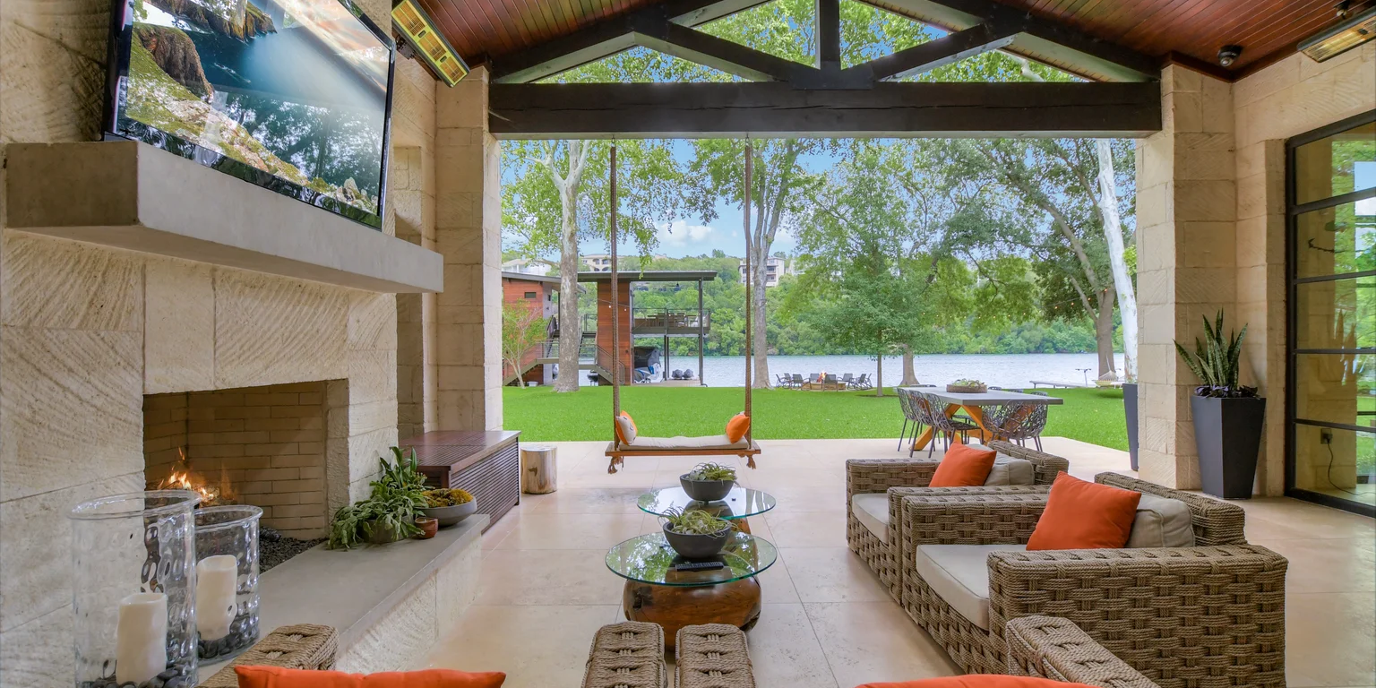 A covered outdoor living area with a fireplace, TV, and wicker furniture overlooking a lake and grassy lawn.