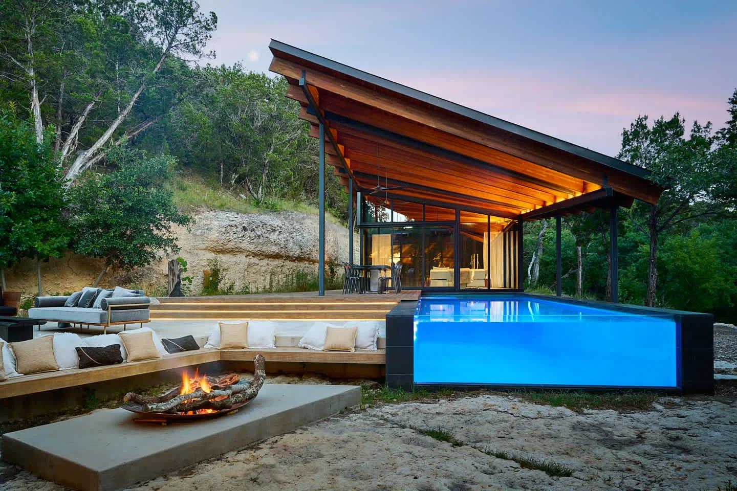 A modern house with a large wooden roof and glass walls next to a glowing blue pool and an outdoor fire pit seating area at dusk.​​​​‌﻿‍﻿​‍​‍‌‍﻿﻿‌﻿​‍‌‍‍‌‌‍‌﻿‌‍‍‌‌‍﻿‍​‍​‍​﻿‍‍​‍​‍‌﻿​﻿‌‍​‌‌‍﻿‍‌‍‍‌‌﻿‌​‌﻿‍‌​‍﻿‍‌‍‍‌‌‍﻿﻿​‍​‍​‍﻿​​‍​‍‌‍‍​‌﻿​‍‌‍‌‌‌‍‌‍​‍​‍​﻿‍‍​‍​‍​‍﻿﻿‌﻿​﻿‌﻿‌​‌﻿‌‌‌‍‌​‌‍‍‌‌‍﻿﻿​‍﻿﻿‌‍‍‌‌‍﻿‍‌﻿‌​‌‍‌‌‌‍﻿‍‌﻿‌​​‍﻿﻿‌‍‌‌‌‍‌​‌‍‍‌‌﻿‌​​‍﻿﻿‌‍﻿‌‌‍﻿﻿‌‍‌​‌‍‌‌​﻿﻿‌‌﻿​​‌﻿​‍‌‍‌‌‌﻿​﻿‌‍‌‌‌‍﻿‍‌﻿‌​‌‍​‌‌﻿‌​‌‍‍‌‌‍﻿﻿‌‍﻿‍​﻿‍﻿‌‍‍‌‌‍‌​​﻿﻿‌​﻿‌﻿‌‍​‌‌‍​‍​﻿‌﻿​﻿​‌​﻿​﻿​﻿‌‍​﻿​﻿​‍﻿‌‌‍​‍​﻿‌‌​﻿​​​﻿​‍​‍﻿‌​﻿‌​‌‍​‍‌‍​‍​﻿​‍​‍﻿‌‌‍​‍​﻿​‍​﻿​﻿​﻿​​​‍﻿‌​﻿‍‌​﻿‌​‌‍​‍​﻿​​​﻿‍​‌‍‌‌‌‍‌‍​﻿​‌​﻿‍​​﻿‍​‌‍‌‍​﻿‍‌​﻿‍﻿‌﻿‌​‌﻿‍‌‌﻿​​‌‍‌‌​﻿﻿‌‌﻿​​‌‍﻿﻿‌﻿​﻿‌﻿‌​​﻿‍﻿‌﻿​​‌‍​‌‌﻿‌​‌‍‍​​﻿﻿‌‌‍​﻿‌‍﻿﻿‌﻿‌‍‌‍‌‌‌﻿​‍‌​‍‌‌‍﻿‌‌‍​‌‌‍‌﻿‌‍‌‌​‍﻿‍‌‍​‌‌‍﻿​‌﻿‌​​﻿﻿﻿‌‍​‍‌‍​‌‌﻿​﻿‌‍‌‌‌‌‌‌‌﻿​‍‌‍﻿​​﻿﻿‌​‍‌‌​﻿​‍‌​‌‍‌﻿​﻿‌﻿‌​‌﻿‌‌‌‍‌​‌‍‍‌‌‍﻿﻿​‍‌‍‌‍‍‌‌‍‌​​﻿﻿‌​﻿‌﻿‌‍​‌‌‍​‍​﻿‌﻿​﻿​‌​﻿​﻿​﻿‌‍​﻿​﻿​‍﻿‌‌‍​‍​﻿‌‌​﻿​​​﻿​‍​‍﻿‌​﻿‌​‌‍​‍‌‍​‍​﻿​‍​‍﻿‌‌‍​‍​﻿​‍​﻿​﻿​﻿​​​‍﻿‌​﻿‍‌​﻿‌​‌‍​‍​﻿​​​﻿‍​‌‍‌‌‌‍‌‍​﻿​‌​﻿‍​​﻿‍​‌‍‌‍​﻿‍‌​‍‌‍‌﻿‌​‌﻿‍‌‌﻿​​‌‍‌‌​﻿﻿‌‌﻿​​‌‍﻿﻿‌﻿​﻿‌﻿‌​​‍‌‍‌﻿​​‌‍​‌‌﻿‌​‌‍‍​​﻿﻿‌‌‍​﻿‌‍﻿﻿‌﻿‌‍‌‍‌‌‌﻿​‍‌​‍‌‌‍﻿‌‌‍​‌‌‍‌﻿‌‍‌‌​‍﻿‍‌‍​‌‌‍﻿​‌﻿‌​​‍‌‍‌﻿​​‌‍‌‌‌﻿​‍‌﻿​﻿‌﻿​​‌‍‌‌‌‍​﻿‌﻿‌​‌‍‍‌‌﻿‌‍‌‍‌‌​﻿﻿‌‌﻿​​‌﻿‌‌‌‍​‍‌‍﻿​‌‍‍‌‌﻿​﻿‌‍‍​‌‍‌‌‌‍‌​​‍​‍‌﻿﻿‌