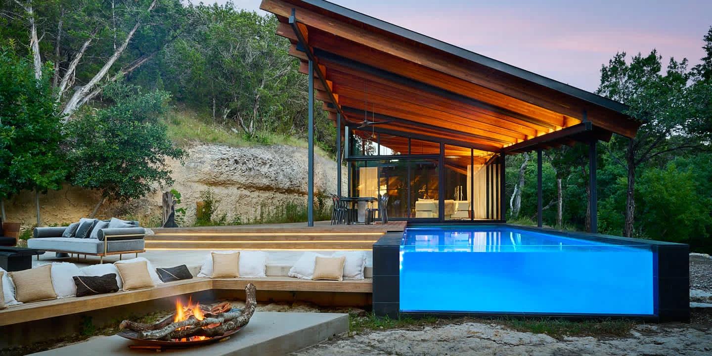 A modern house with a large wooden roof and glass walls next to a glowing blue pool and an outdoor fire pit seating area at dusk.