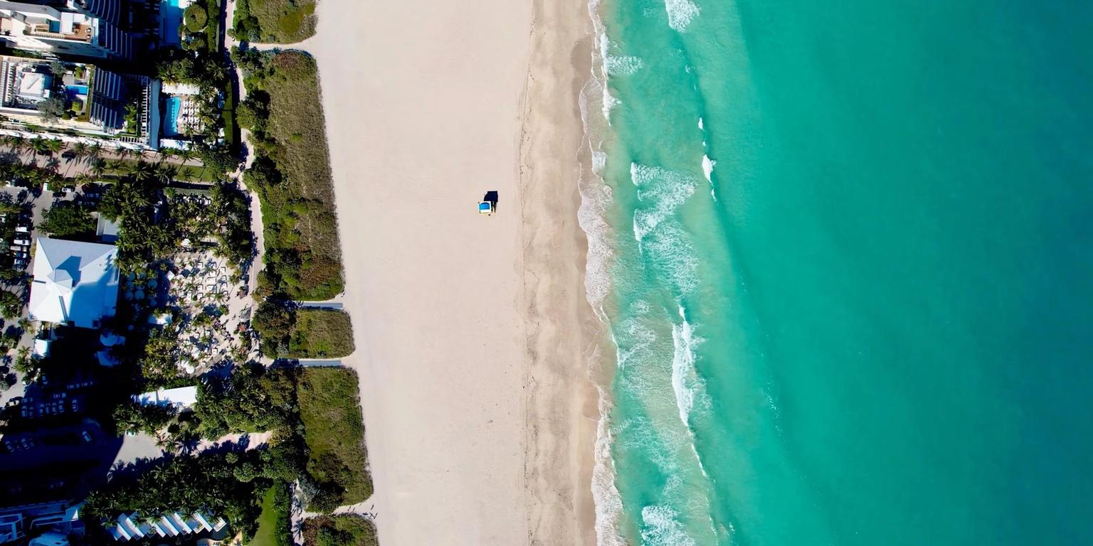 Aerial view of a white sand beach, turquoise ocean with waves, a single beach umbrella, and a coastal city.