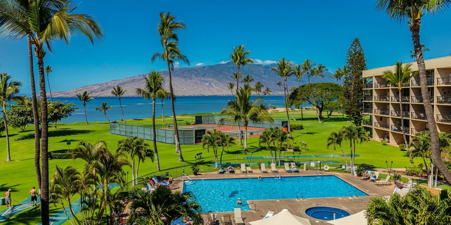 A tropical resort landscape with a swimming pool, hotel, palm trees, green lawns, tennis courts, ocean, and distant mountains under a clear blue sky.