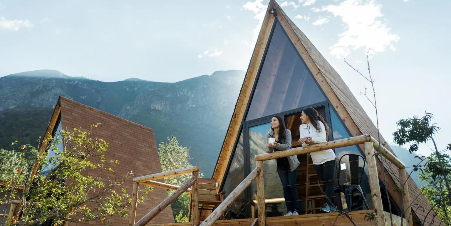 Two women stand on the balcony of a modern A-frame cabin with mountains in the background.