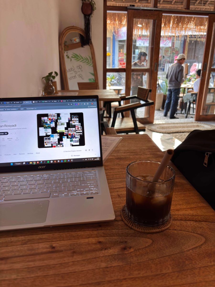 A cozy café workspace with a silver laptop open on a portfolio website beside a glass of iced coffee on a wooden table. Warm natural light streams through large wooden-framed doors, while customers sit and walk outside, creating a relaxed atmosphere ideal for remote work.
