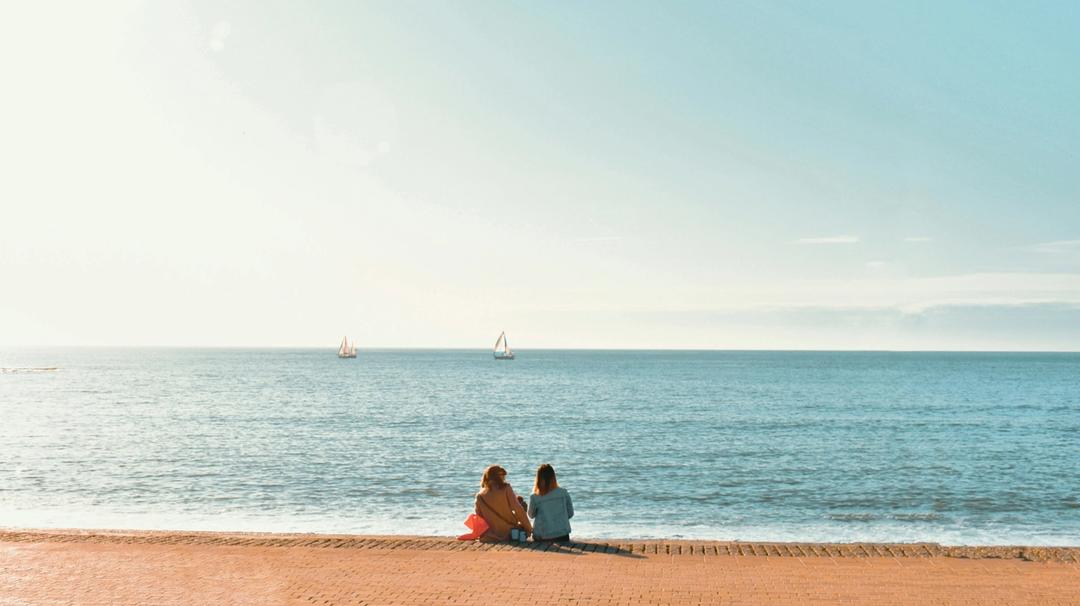 Two women sitting and facing the ocean during daytime.