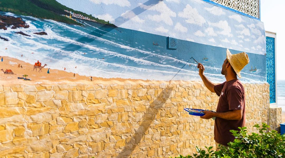 Person painting a seaside landscape mural on a building wall.