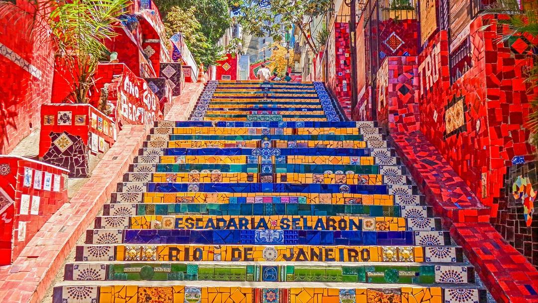 Colorful mosaic-tiled staircase in Rio de Janeiro, lined with vibrant red walls, artwork, and tropical plants.